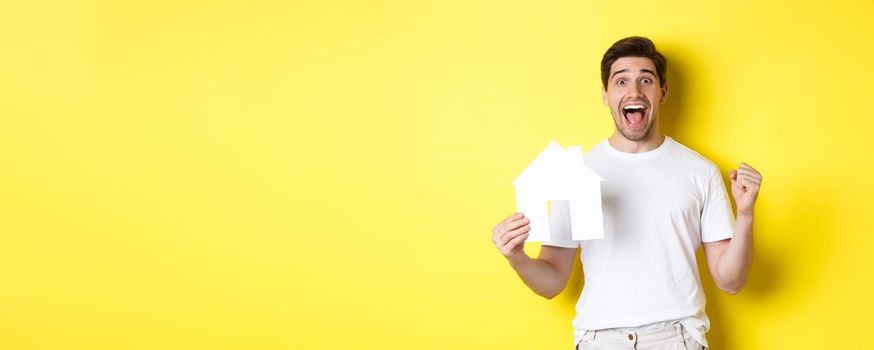 Real estate concept. Excited man holding paper house model and celebrating, standing happy over yellow background.