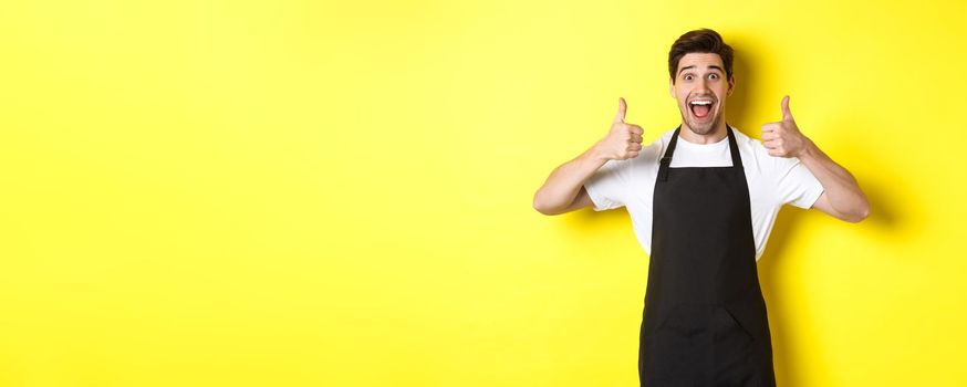 Cheerful man barista in black apron showing thumbs-up, recommending cafe or restaurant, standing against yellow background.