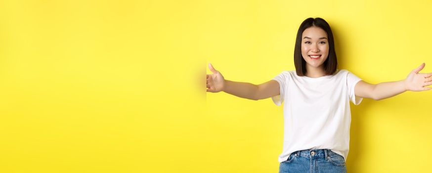 Beauty and fashion concept. Friendly asian woman spread out hands and smiling, waiting for hugs, inviting you, welcome someone and looking happy, standing over yellow background.