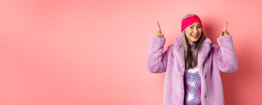 Stylish asian woman smiling happy, pointing fingers up and showing advertisement, wearing winter fake-fur coat and headband, pink background.
