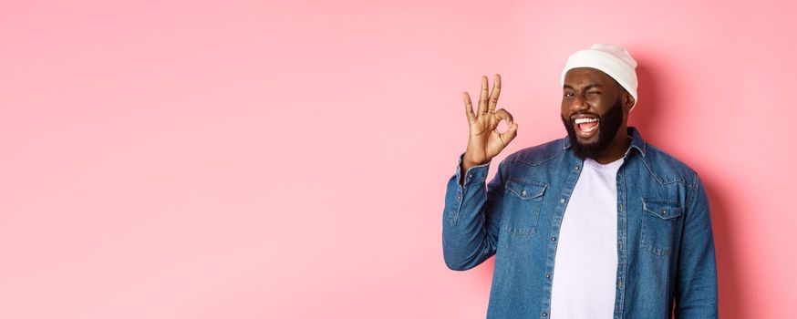 Happy smiling african american man showing okay sign, approve and praise good offer, standing over pink background.