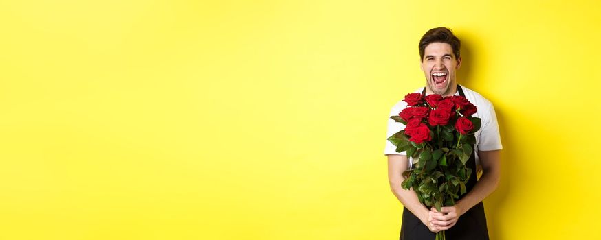 Funny young salesman in black apron holding bouquet of roses, florist laughing and standing over yellow background.