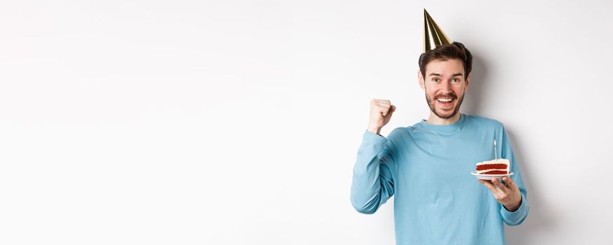 Celebration and holidays concept. Cheerful young man celebrating birthday in party hat, holding bday cake and looking happy, standing on white background.