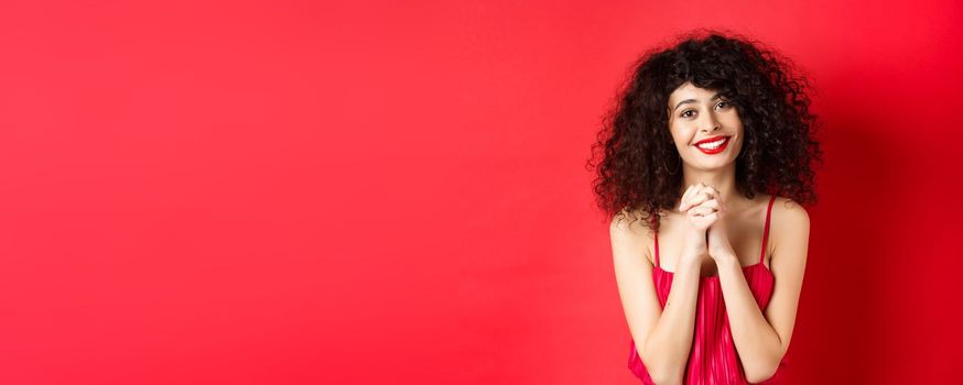 Image of beautiful curly-haired woman in red dress and makeup, saying thank you, looking grateful and smiling happy at camera, studio background.