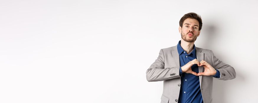 Romantic man in suit showing heart sign and pucker lips for kiss, express love and passion, like his lover, standing on white background.