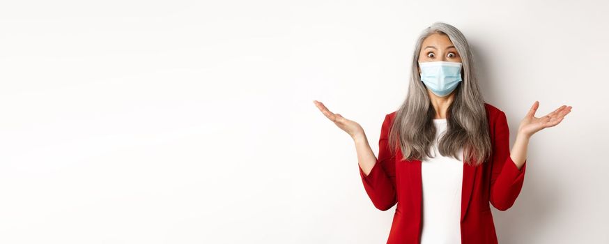 Covid-19 and business people concept. Asian businesswoman in face mask looking surprised, staring at camera in awe, standing over white background.