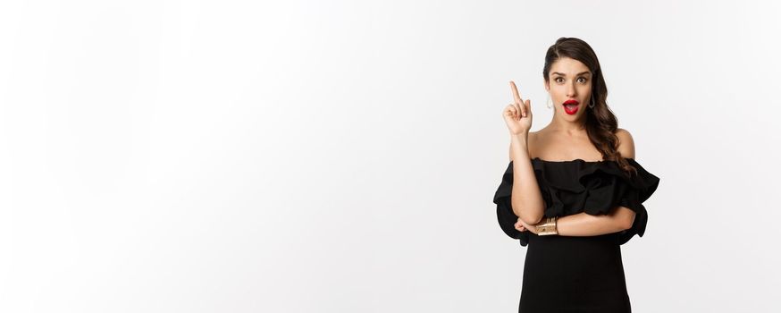 Fashion and beauty. Attractive caucasian woman in black dress having an idea, raising finger and saying suggestion, white background.