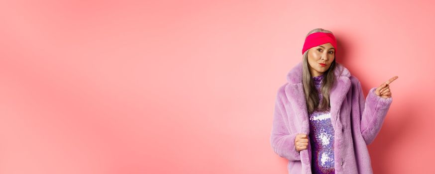 Trendy asian senior woman in cool purple winter coat pointing finger right, showing advertisement and looking sassy at camera, pink background.
