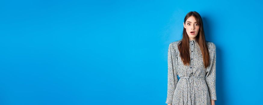 Shocked cute girl in dress looking at camera with opened mouth, gasping startled, see something shocking, standing on blue background.