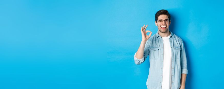 Confident smiling man in glasses showing ok sign, winking to guarantee or recommend something, blue background.