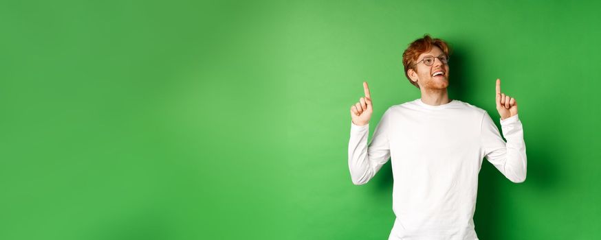 Cheerful handsome man in glasses checking out sale deal, pointing and looking up with happy and dreamy smile, standing over green background.