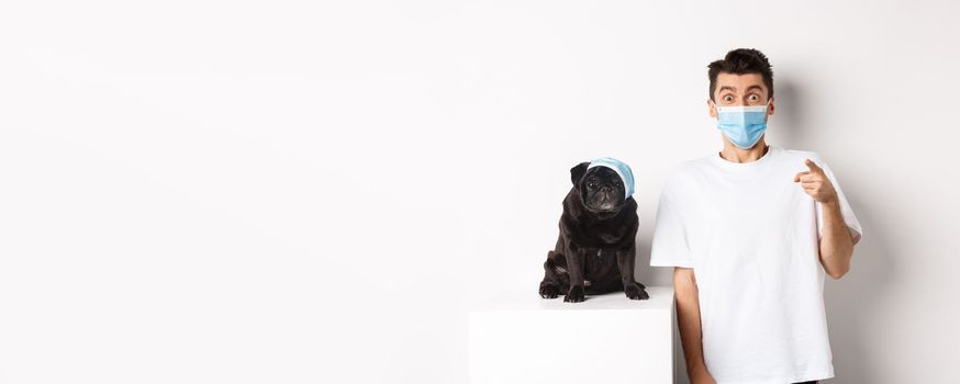 Covid-19, animals and quarantine concept. Happy dog owner and cute pug wearing medical masks, man pointing finger at camera amazed, white background.