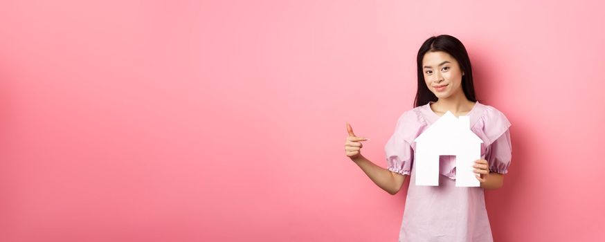 Real estate and insurance concept. Beautiful asian woman pointing at paper house cutout, showing agency logo, standing in dress on pink background.