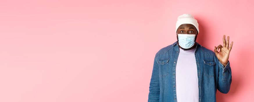 Coronavirus, lifestyle and social distancing concept. Impressed african-american man in face mask, showing okay sign, like and agree, standing over pink background.