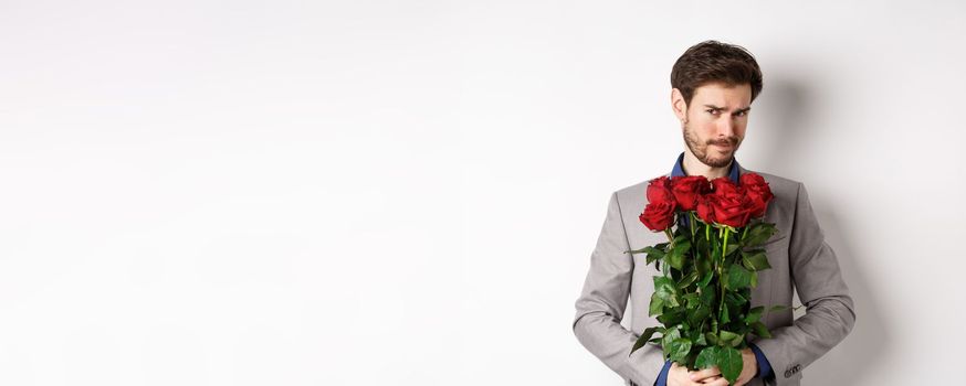 Indecisive bearded man in suit, holding flowers roses and looking at camera unsure, going on romantic date, standing over white background.