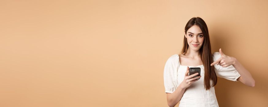 Attractive woman smiling and pointing at smartphone, showing online offer on phone, standing on beige background.