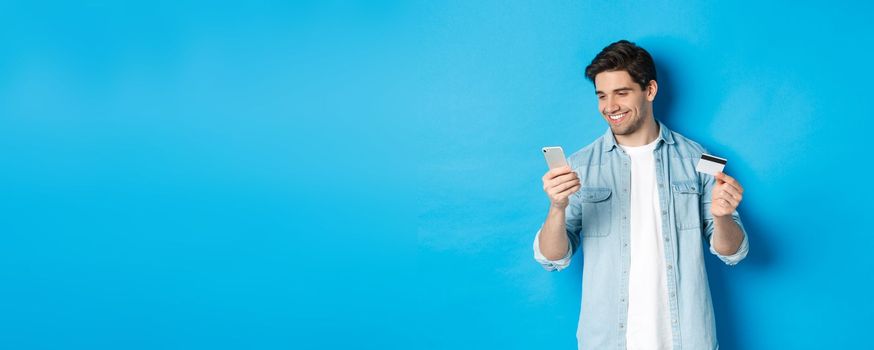 Young man shopping online with mobile application, holding smartphone and credit card, standing over blue background.