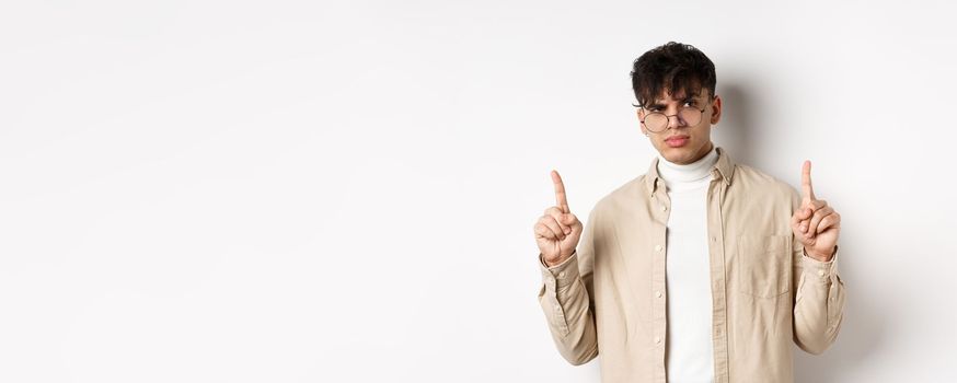 Portrait of young male model in glasses looking suspicious and frowning, pointing fingers up with disbelief, standing on white background.