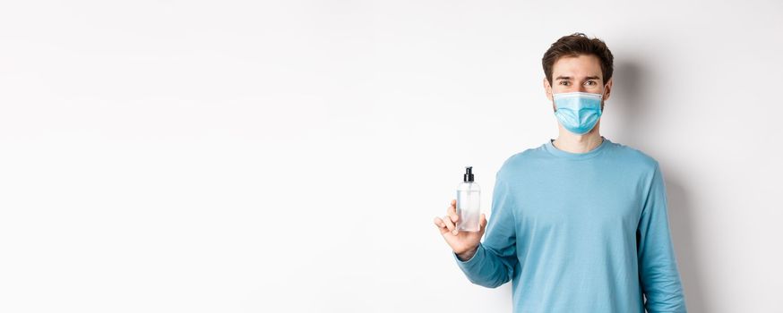 Covid-19, health and quarantine concept. Portrait of smiling man in medical mask showing hand sanitizer bottle, standing over white background.
