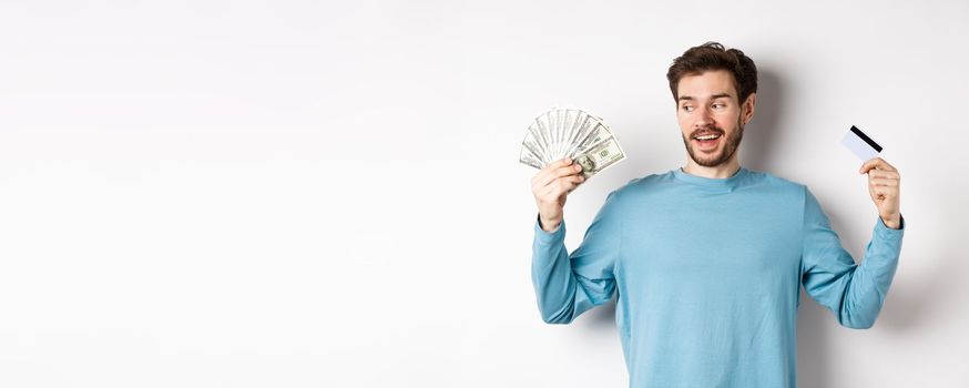 Excited bearded man choosing between money and plastic credit card, payment with cash or contactless, standing over white background.