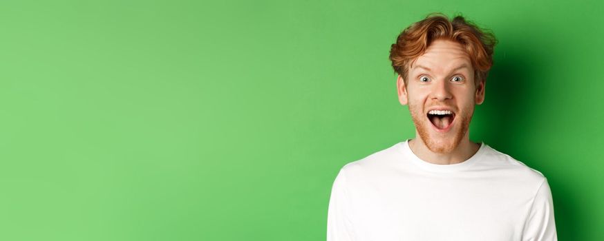 Emotions and fashion concept. Close up of surprised redhead man hear amazing news, staring at camera joyful, standing over green background.