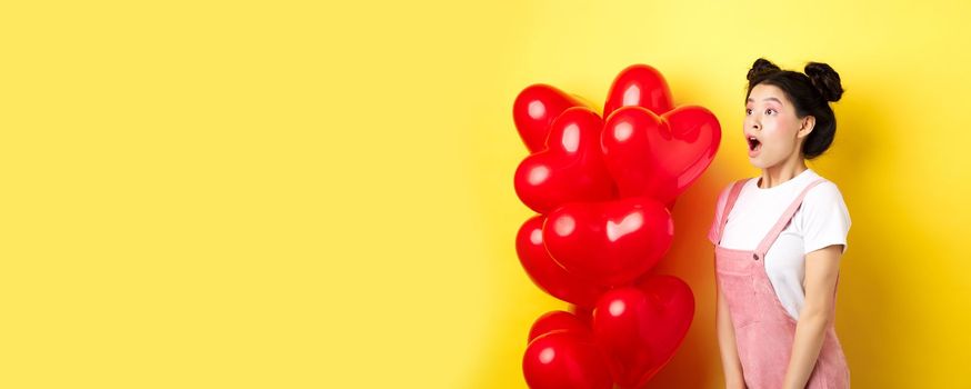 Valentines day and relationship concept. Fashion girl looking amazed, gasping and say wow, staring left while standing near red hearts balloons, surprise from lover, yellow background.