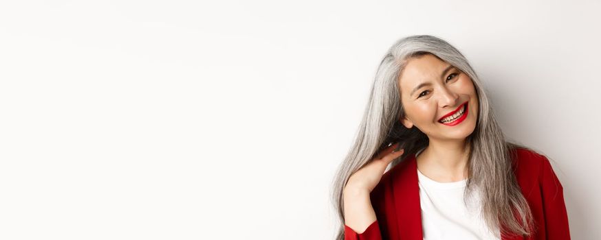 Beauty and aging concept. Close up of asian senior woman with red lips, long healthy grey hair, smiling at camera, standing over white background.