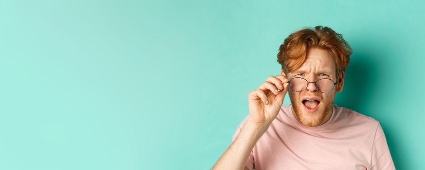 Close up portrait of redhead guy take-off glasses and looking confused at something strange, standing shocked over turquoise background.