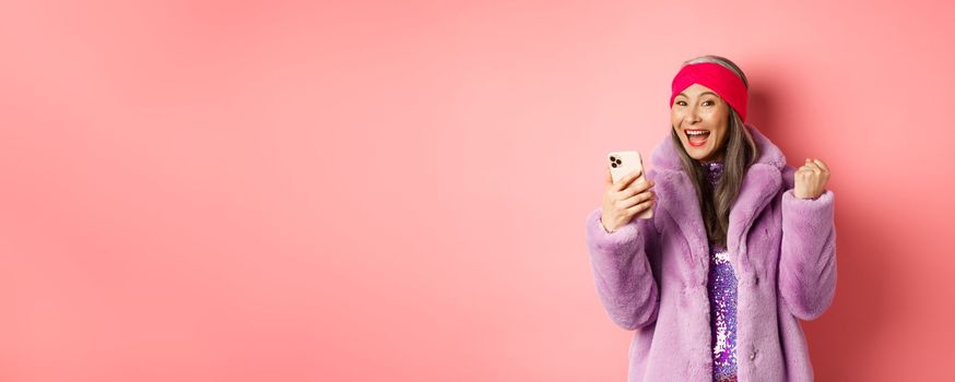 Online shopping and fashion concept. Happy asian senior woman winning prize in internet, holding mobile phone and making fist pump, scream of joy, standing over pink background.