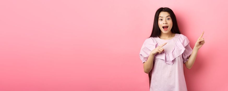 Excited asian teenage girl pointing fingers right, smiling happy and amused, showing cool news, standing on pink background.