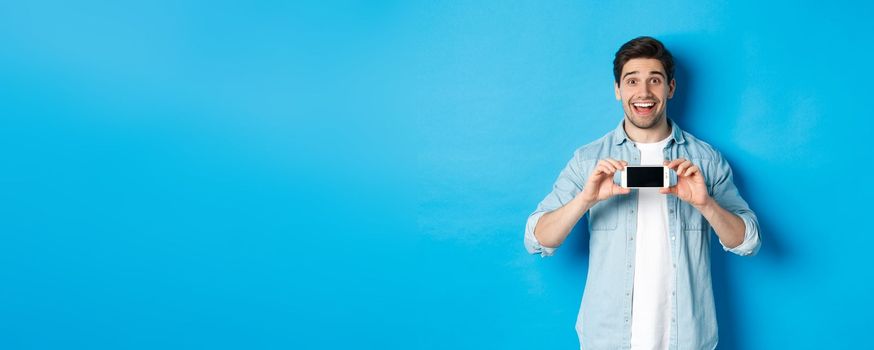 Amazed smiling man showing smartphone screen, internet promo offer, standing against blue background.