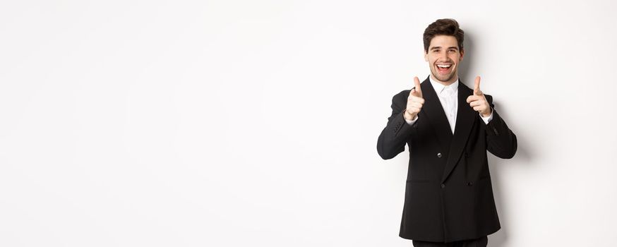 Concept of new year party, celebration and lifestyle. Handsome and successful guy in black suit, pointing fingers at camera and congratulating you, standing over white background.