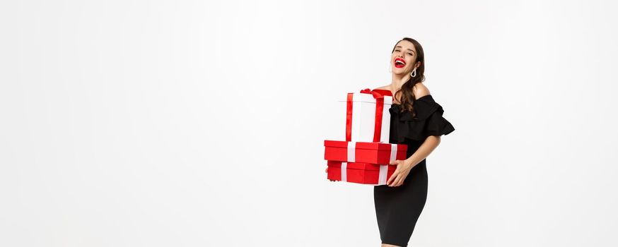 Merry christmas and new year holidays concept. Full length of attractive woman in elegant dress laughing, holding xmas gifts, laughing happy, white background.