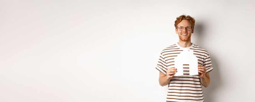 Handsome young man with beard and red hair, wearing glasses and striped t-shirt, showing paper house cutout and smiling, concept of real estate and buying property.