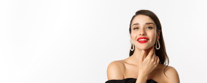 Fashion and beauty concept. Close-up of gorgeous brunette woman with red lips, touching face and smiling self-assured, standing over white background.