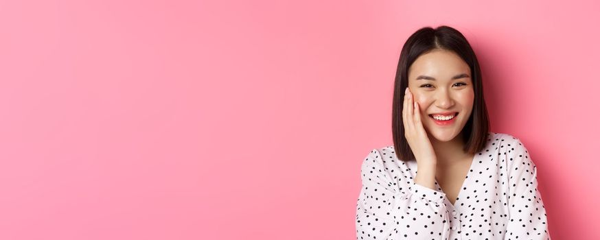 Close-up of adorable asian woman blushing, touching cheek and smiling cute at camera, standing over pink background.