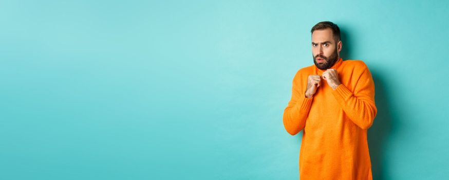 Scared guy jumping startled, looking at something scary, standing in orange sweater, studio background.