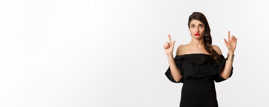 Fashion and beauty. Silly woman in black dress, red lips, looking and pointing fingers up with unamused doubtful expression, white background.