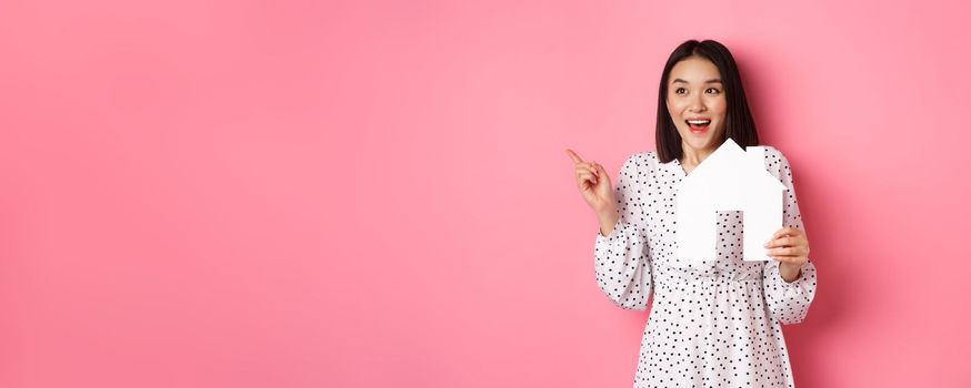 Real estate. Excited asian woman showing paper house model, pointing and looking left at copy space, standing over pink background.