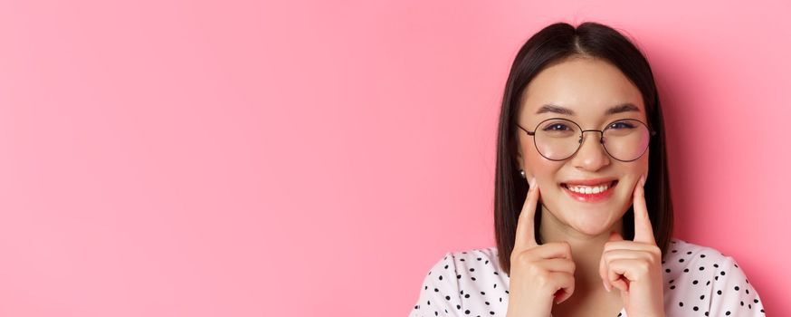 Beauty concept. Headshot of adorable asian girl in trendy glasses smiling, poking cheeks and showing cute dimples, standing over pink background.