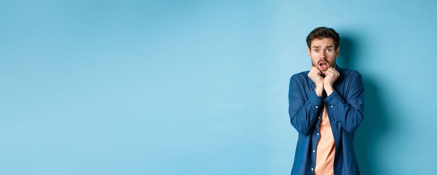 Scared young man gasping and screaming frightened, looking terrified at something, standing on blue background.