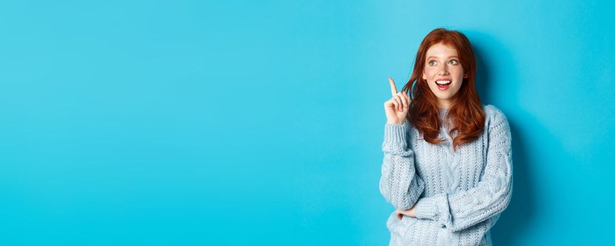 Thoughtful redhead girl having an idea, raising finger and smiling pleased with good plan, standing over blue background.