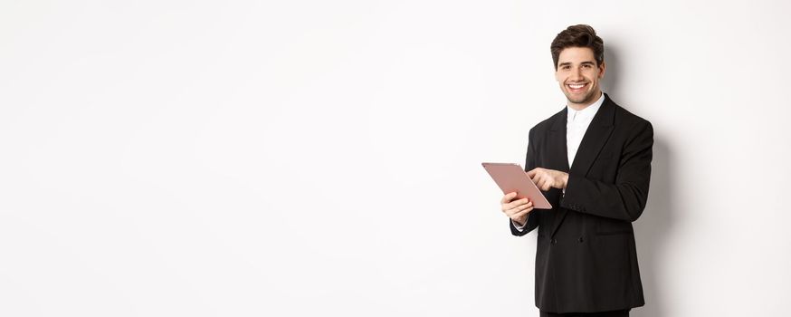Image of handsome businessman in black suit, using digital tablet and smiling, standing against white background.