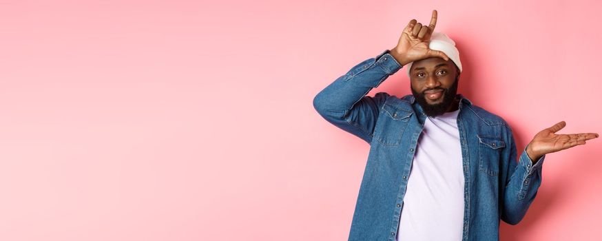 Sad african-american man showing loser sign on forehead and staring at camera, standing over pink background.