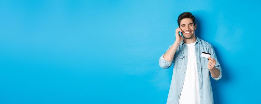 Handsome man calling bank and holding credit card, having mobile conversation, standing over blue background.