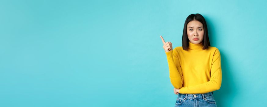 Shopping concept. Sad and gloomy asian girl in yellow sweater pointing finger left, frowning and showing bad news on copy space, standing over blue background.