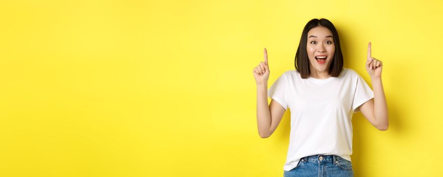 Beauty and fashion concept. Beautiful asian woman in white t-shirt pointing fingers up, standing over yellow background.