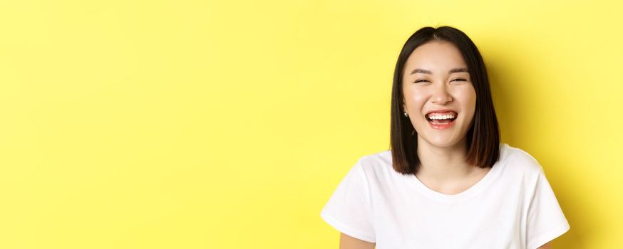 Close up of happy young woman having fun, smiling and laughing carefree, standing in white t-shirt over yellow background.