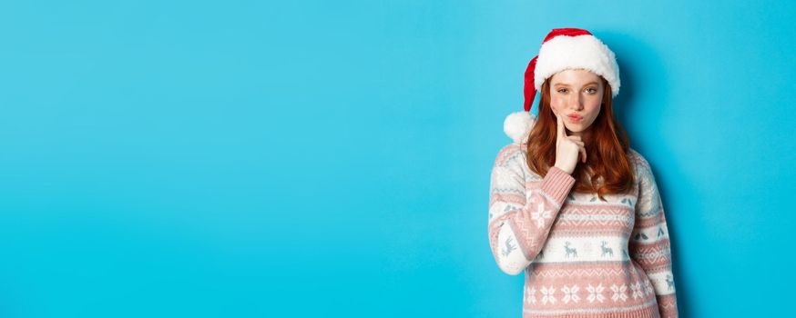 Winter and Christmas Eve concept. Thoughtful redhead teen girl looking at camera, making choice or thinking, wearing santa hat, standing over blue background.