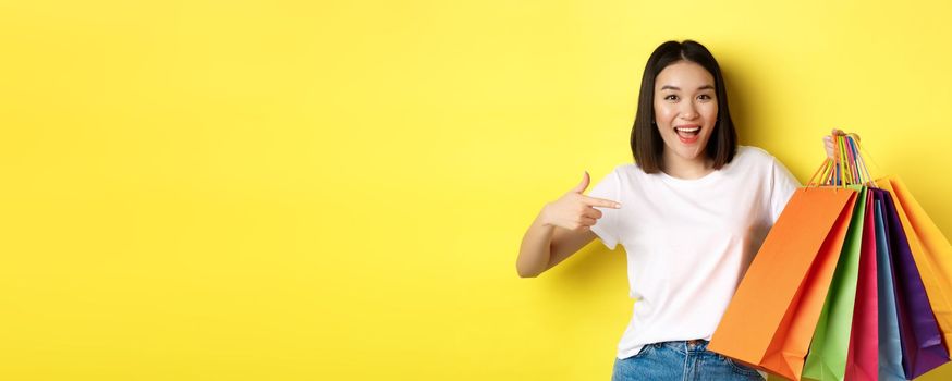 Cheerful asian woman going on shopping, pointing finger at paper bags and smiling, shopper having fun during discounts, yellow background.